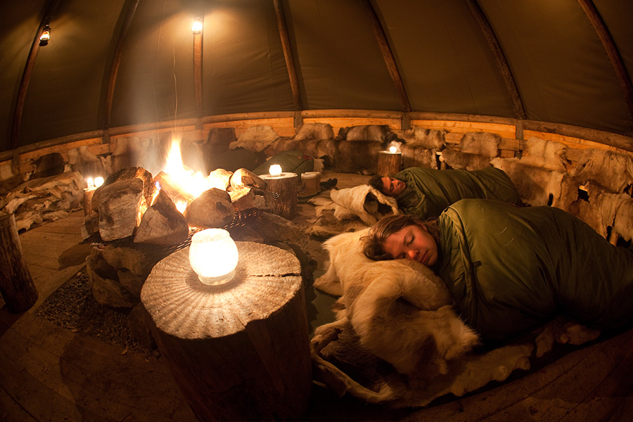 People sleeping on the reindeer skins inside a traditional Sami lavvo tent with the burning fireplace in the middle.