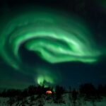 Aurora Borealis forming a round shape above the little hut on top of the hill.