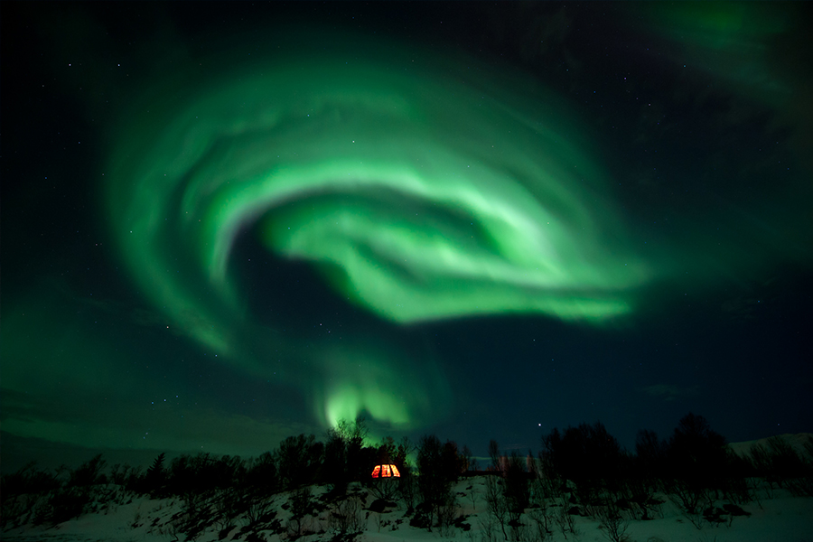 Aurora Borealis forming a round shape above the little hut on top of the hill.