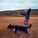 A blond woman smiling and showing thumbs up while hiking in the field with a black dog attached to her waist.