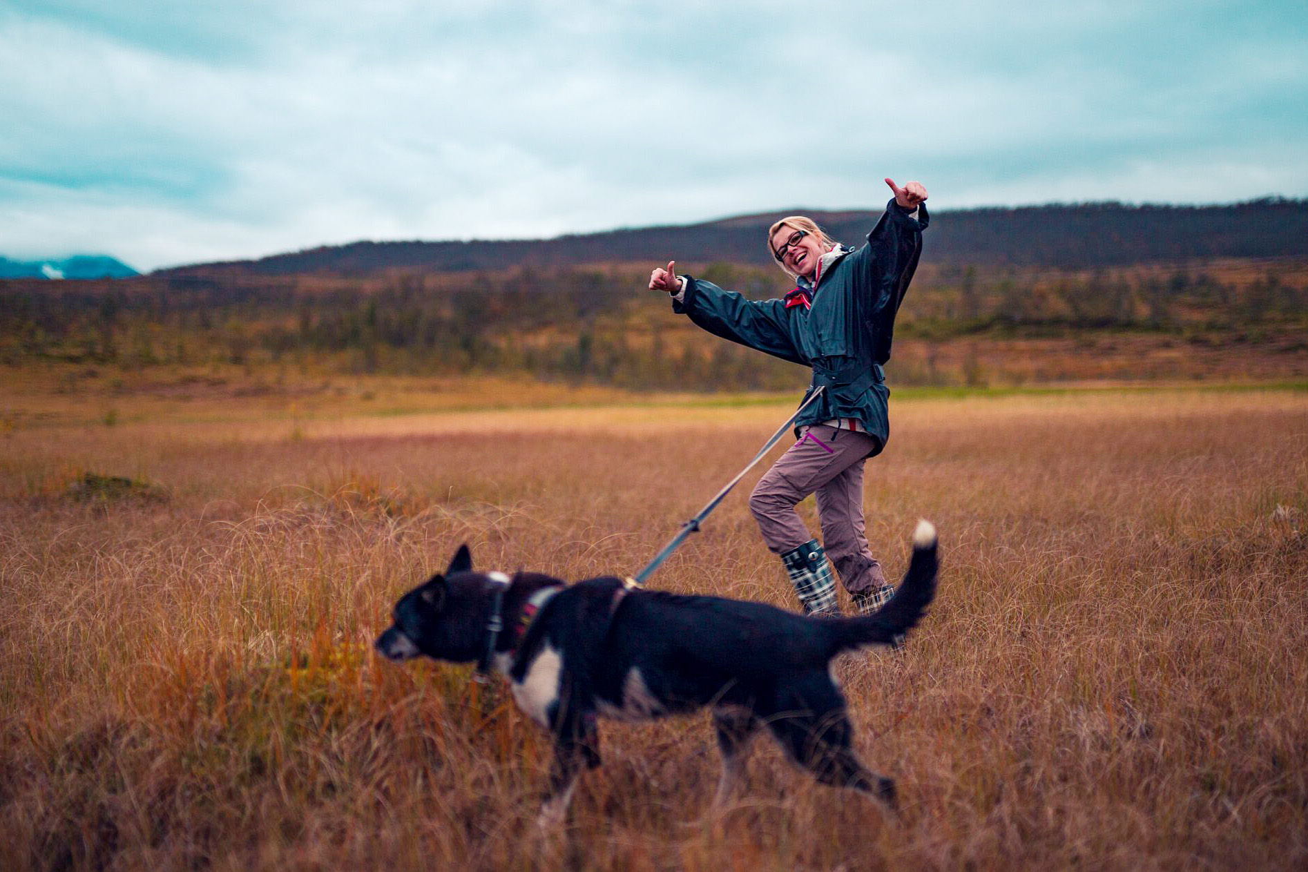 A blond woman smiling and showing thumbs up while hiking in the field with a black dog attached to her waist.