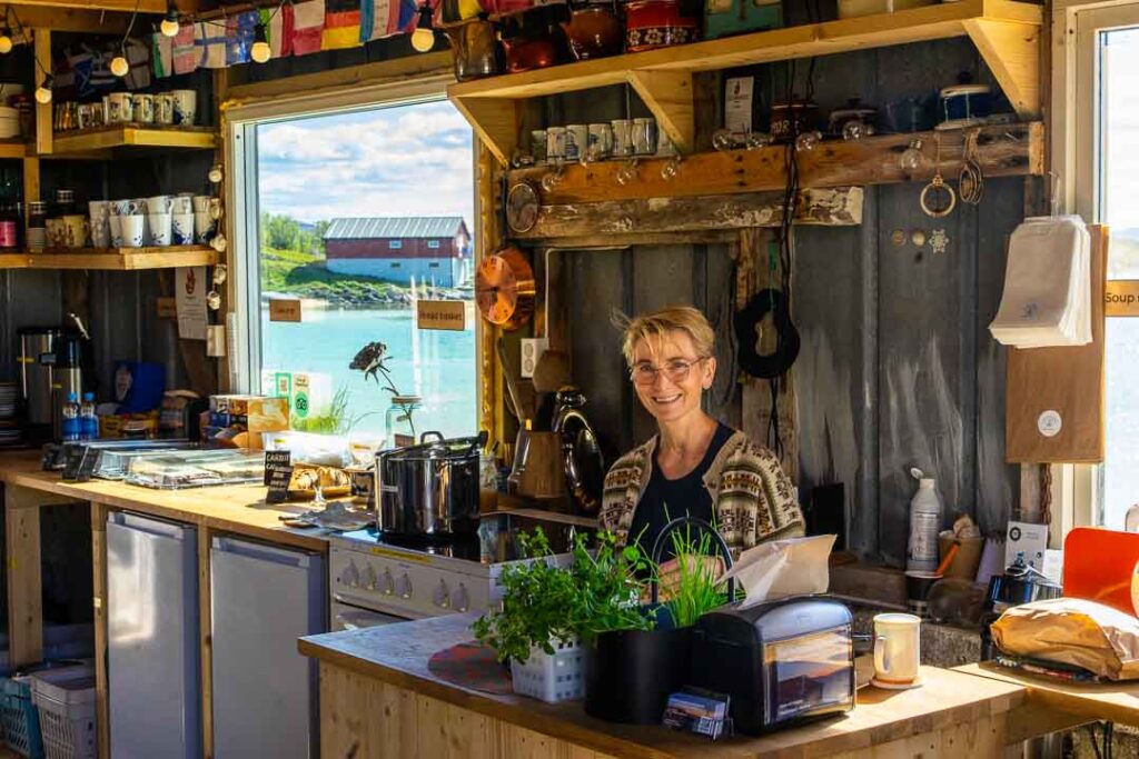 Woman smiling behind the counter of a cozy waterside café, surrounded by wooden shelves, kitchen utensils, and a view of a red building across the water through the window.