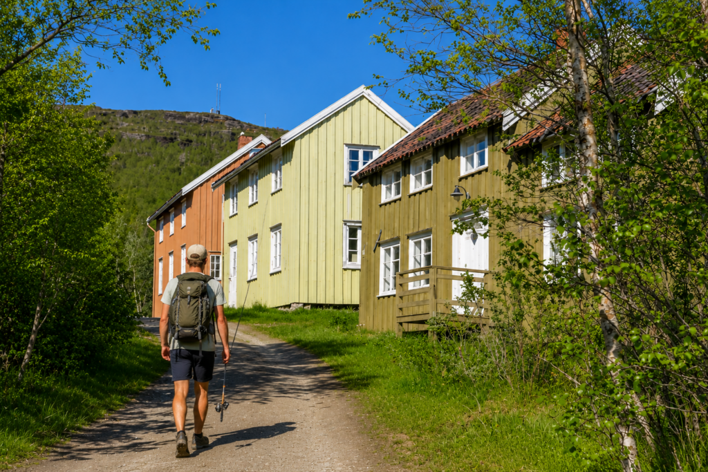 Backpacker walking through a traditional Norwegian village with a fishing rod and backpack on a sunny summer day.