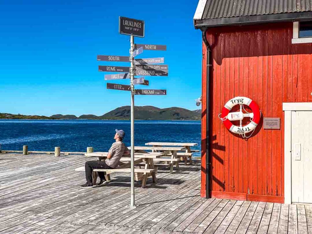 Man relaxing on a bench beside a red waterfront building and directional signpost, overlooking a calm fjord with blue water, distant islands, and clear summer skies in Northern Norway.