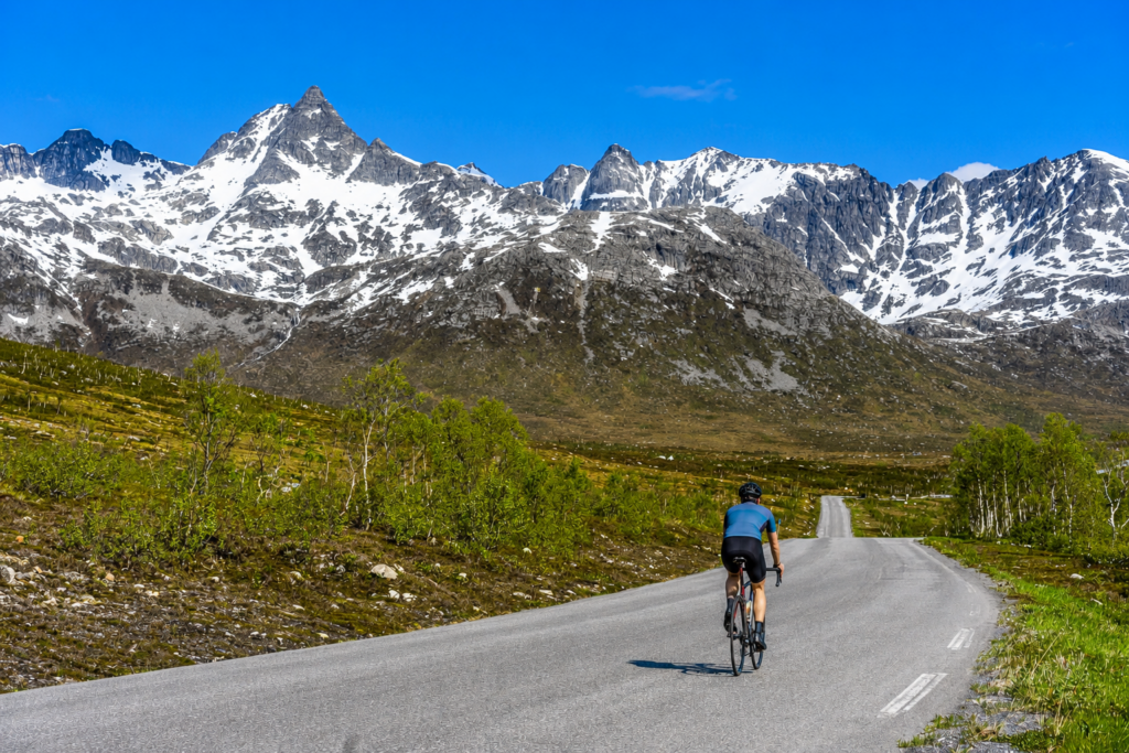 Cyclist riding along a winding mountain road beneath snow-capped peaks in Northern Norway on a clear summer day.