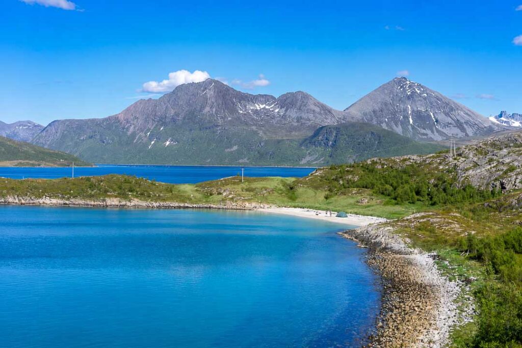 Turquoise bay and white sandy beach surrounded by rugged mountains in Northern Norway, with a small tent, hikers, and a husky visible in the distance on a sunny summer day.
