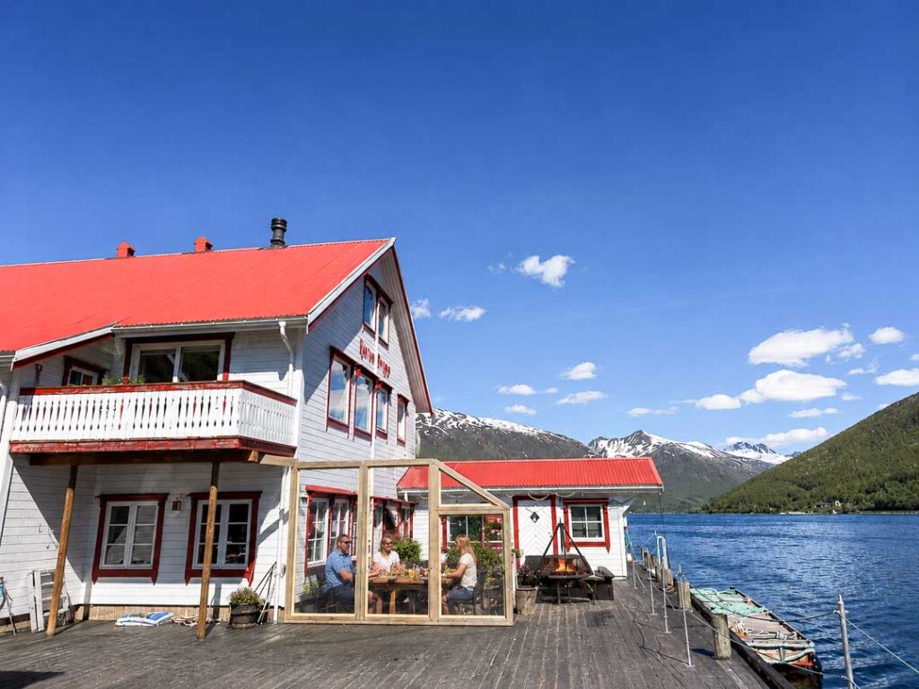 Traditional waterfront lodge with red roofs overlooking a fjord, where three people sit inside a glass-covered terrace beside a small fire, surrounded by mountains, blue water, and a sunny summer sky.